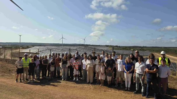 Grupo de pessoas posando para uma foto em frente a um parque de instalação solar e alguns moinhos de vento